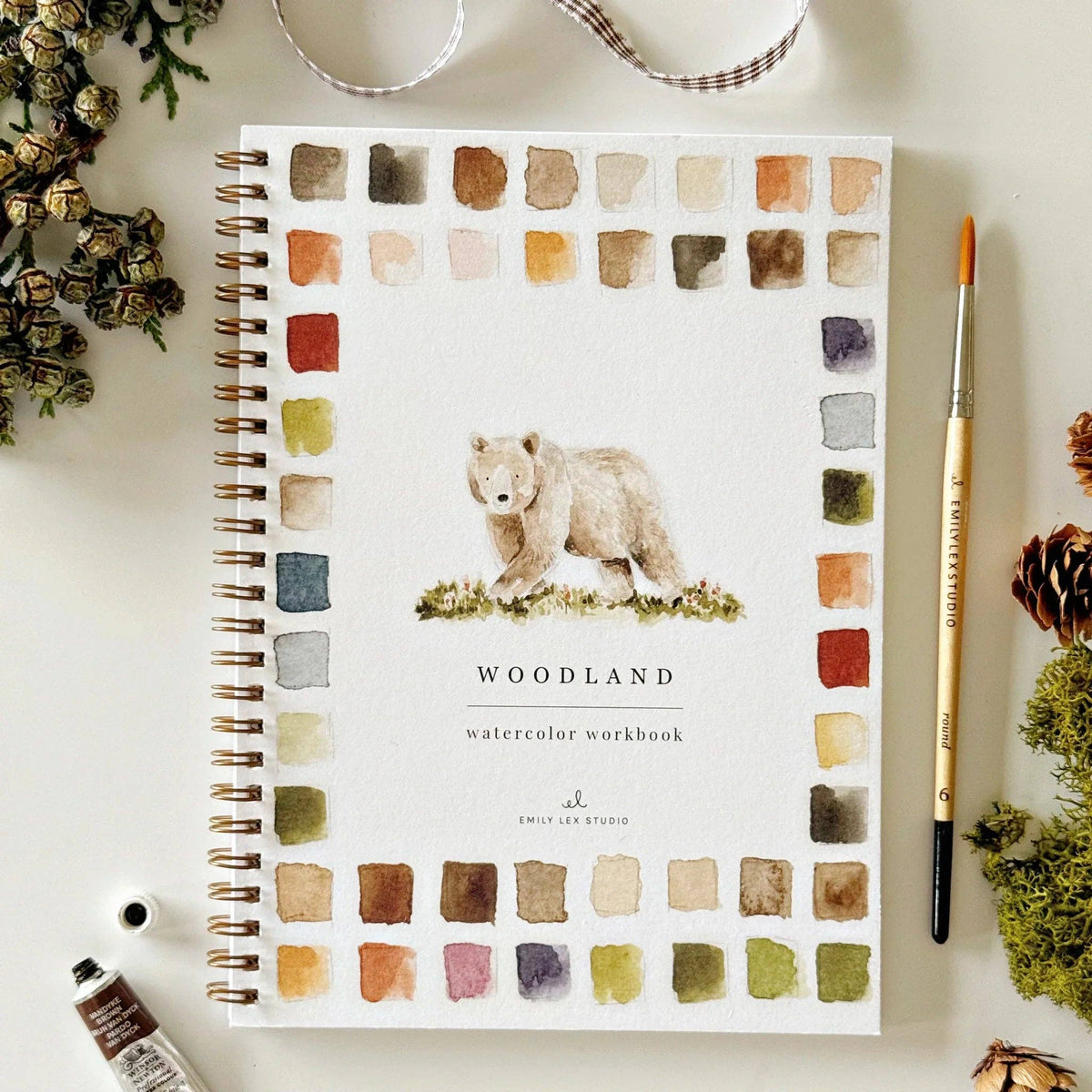 A spiral-bound watercolor workbook titled 'WOODLAND' with a bear illustration and watercolor swatches around the cover, styled on a light surface with a paintbrush, paint tube, and dried botanicals.
