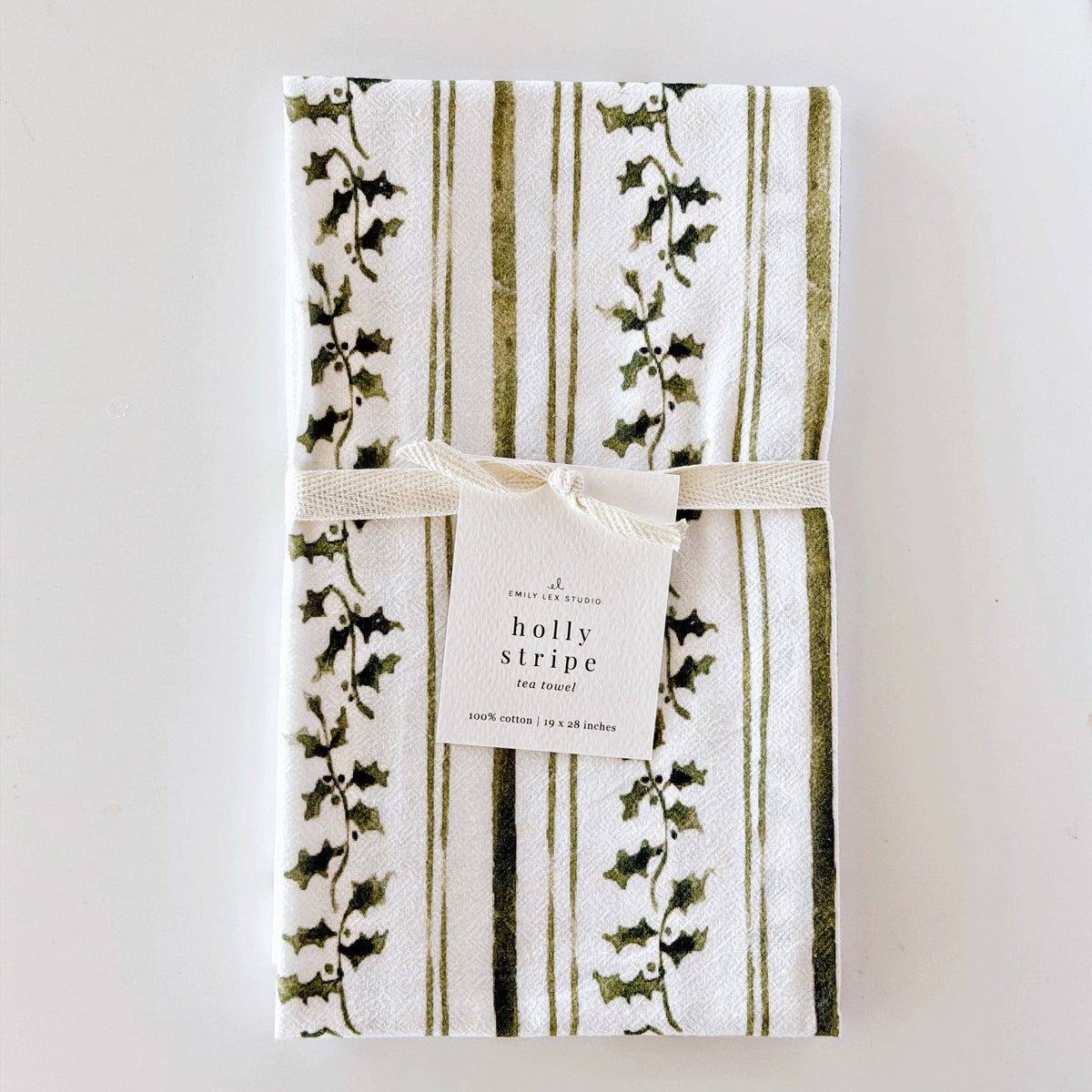 A folded cotton tea towel with a white background, featuring vertical olive green stripes and green holly leaf vines, tied with a cream ribbon and a paper label that reads 'holly stripe tea towel'.
