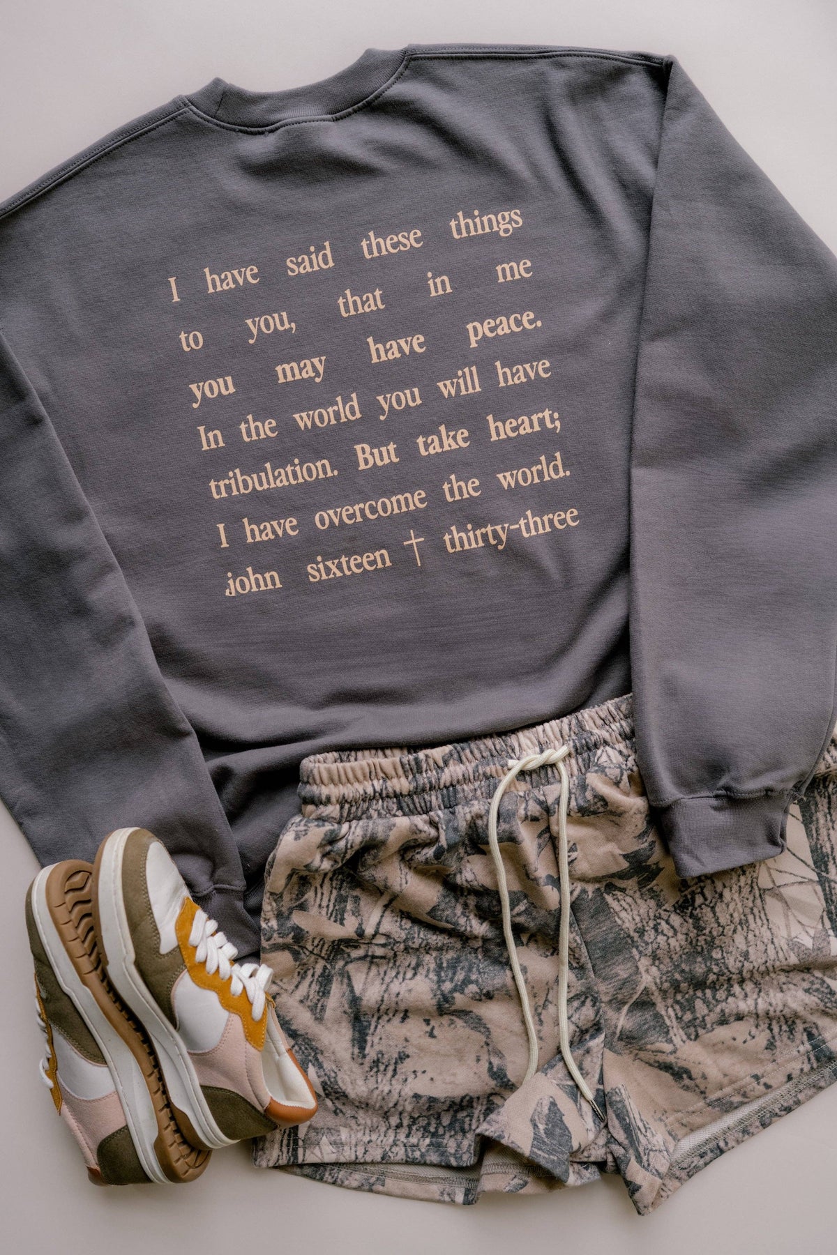 A styled flat-lay of a dark gray crewneck sweatshirt featuring a beige typographic quote on the back, paired with beige and gray patterned shorts and a pair of multicolor sneakers on a light gray background.
