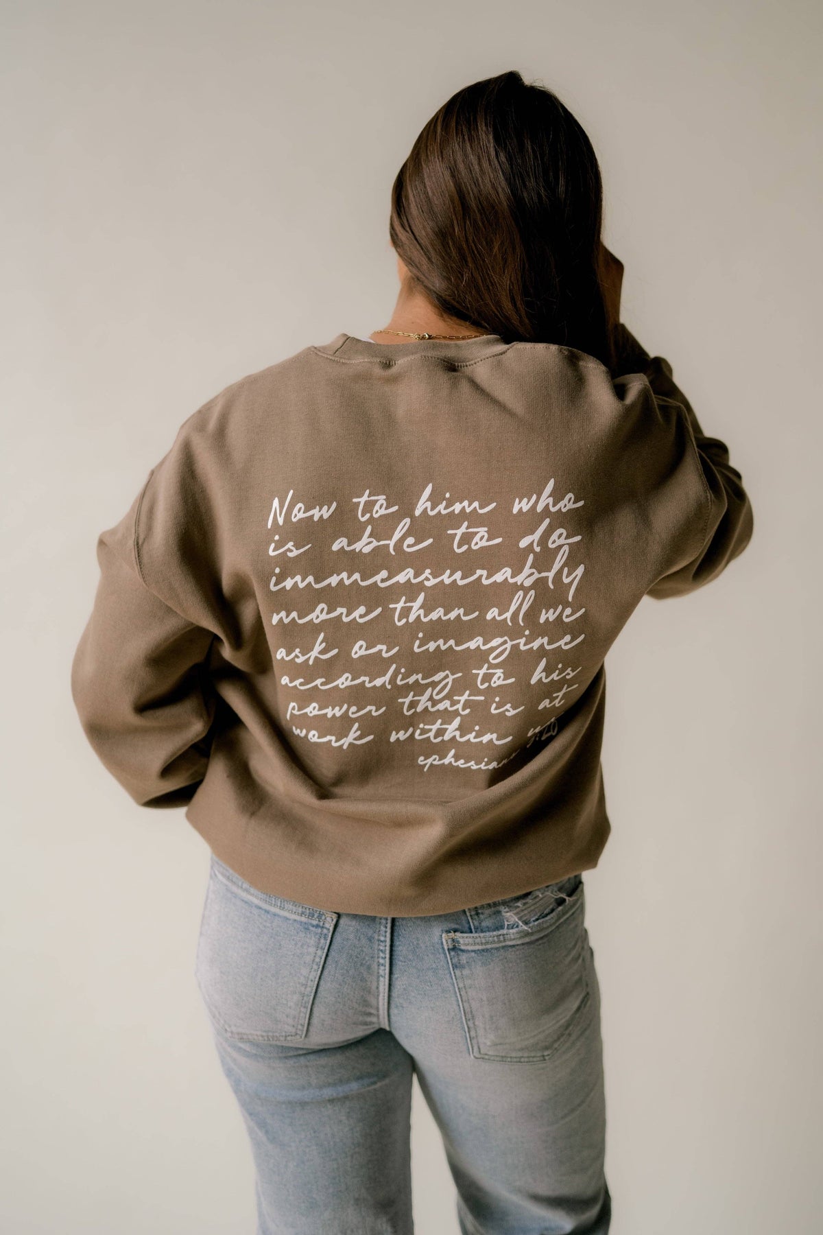 A woman viewed from behind wearing a brown sweatshirt with white cursive text printed on the back, paired with light blue jeans, against a plain light gray background.