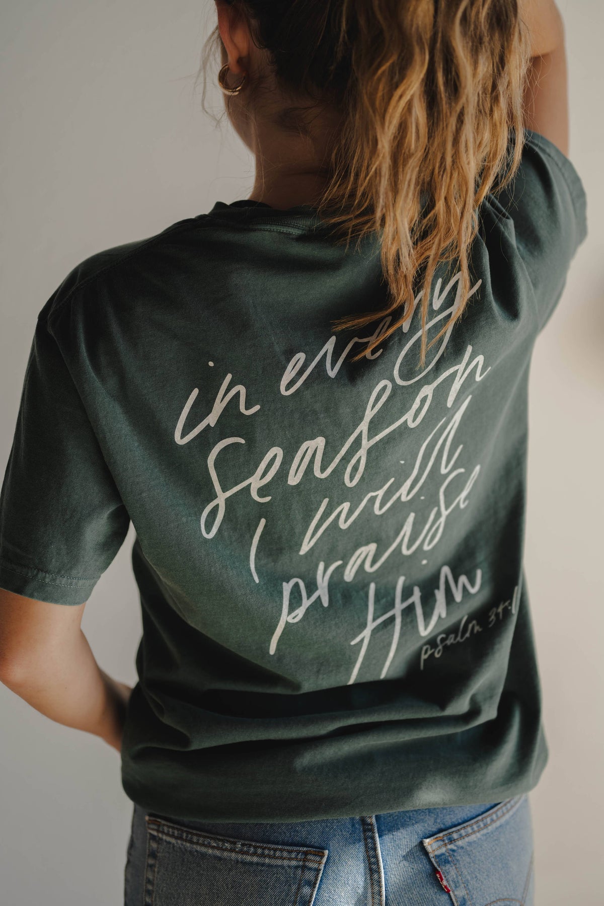 Person wearing a green "in every season" t-shirt with text on the back, standing against a neutral background