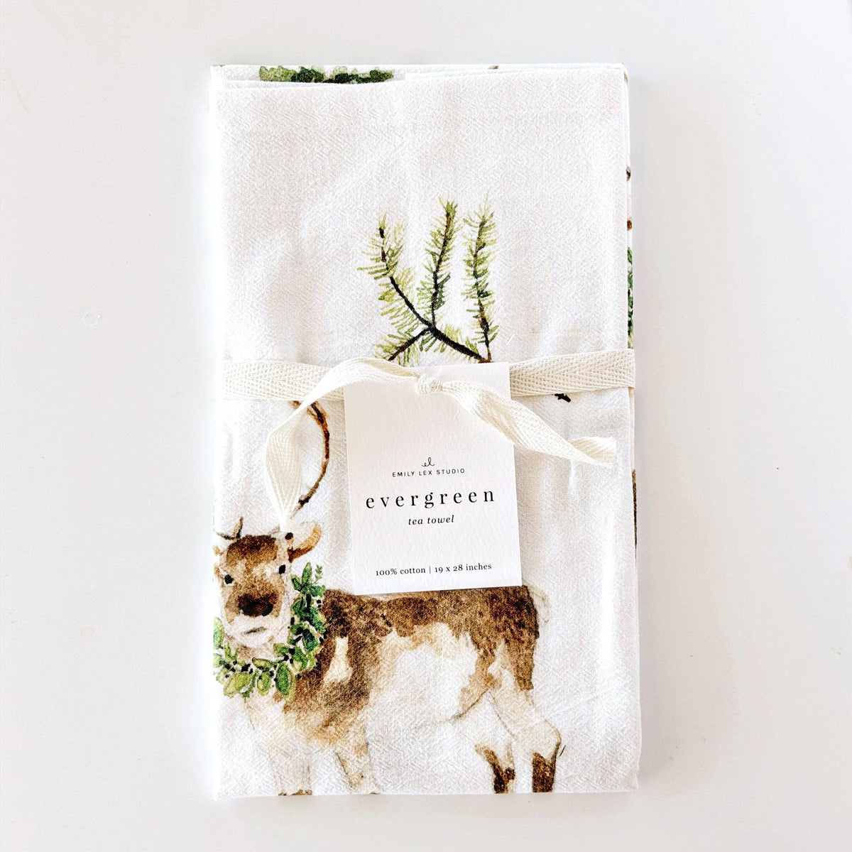 A folded white cotton tea towel with a Christmas deer and pine branch print, tied with a cream ribbon and a paper tag that reads 'evergreen tea towel' on a white background.