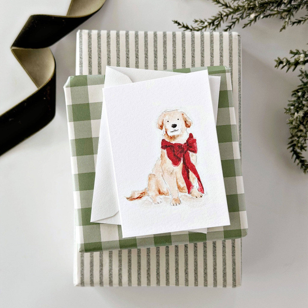A square greeting card featuring a watercolor illustration of a golden retriever puppy wearing a red bow, displayed on top of a green and white striped gift box with a white envelope.