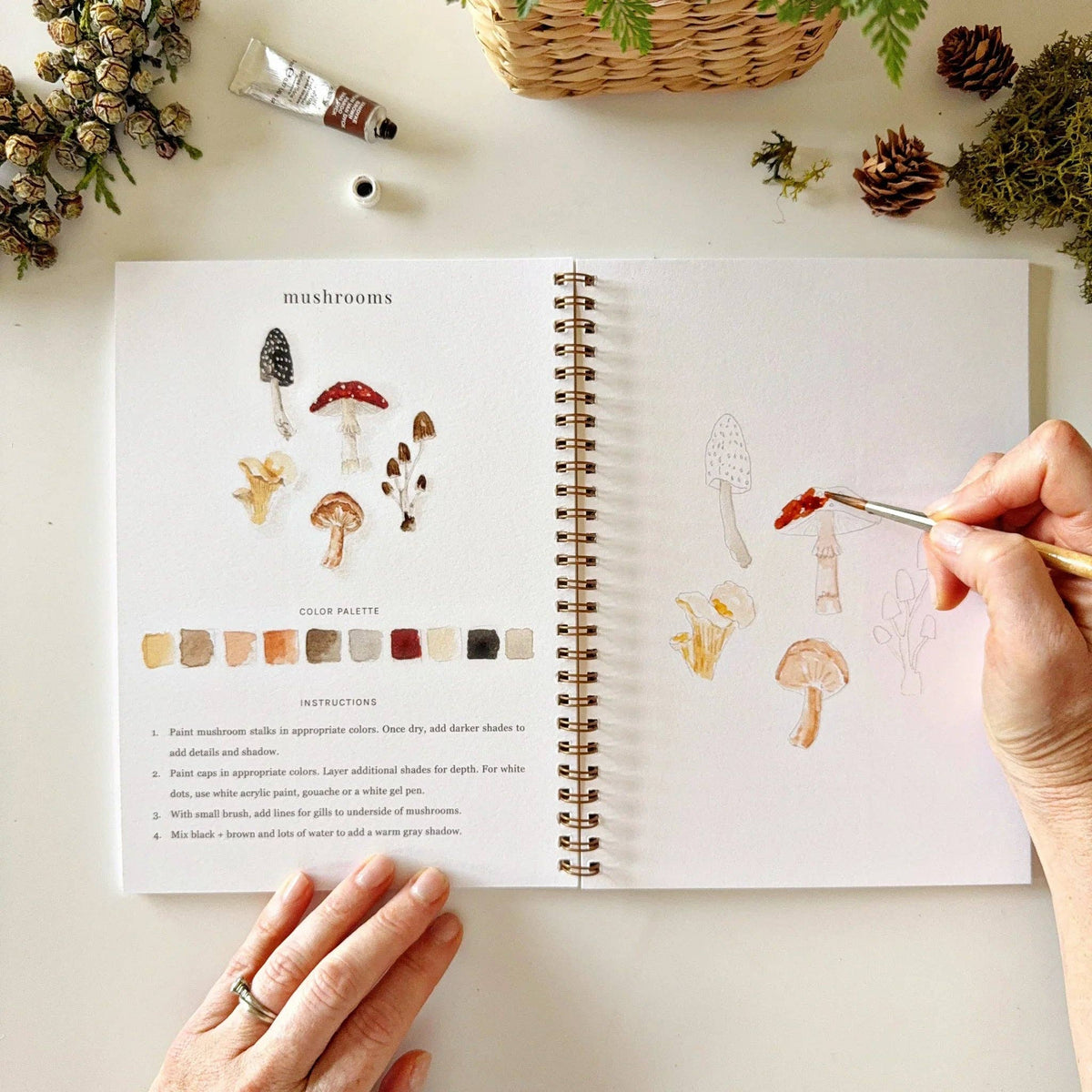 Open book with mushroom illustrations and a hand holding a paintbrush on a light surface.