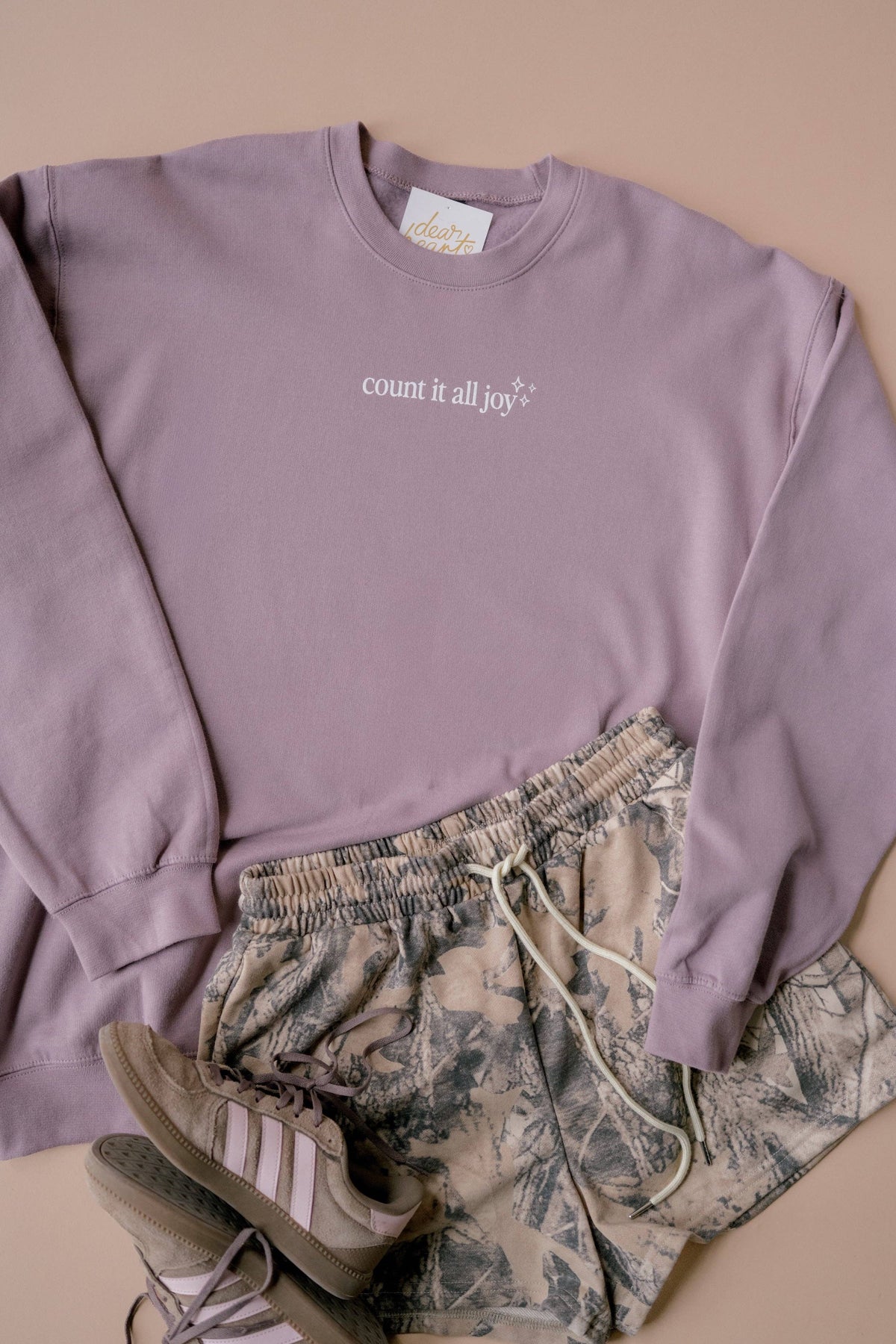 A styled flat-lay of a lavender crewneck sweatshirt with the text 'count it all joy✨' on the chest, paired with camouflage print shorts and a pair of beige sneakers on a solid beige background.