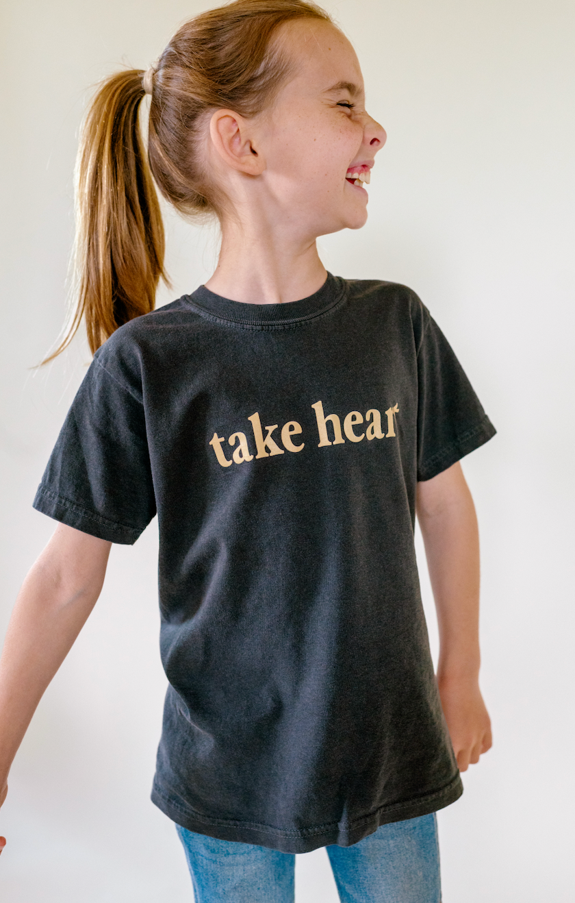 A young girl with a ponytail wearing a dark charcoal gray t-shirt with the words 'take heart' printed in a light beige serif font across the chest, paired with blue jeans against a plain white background.