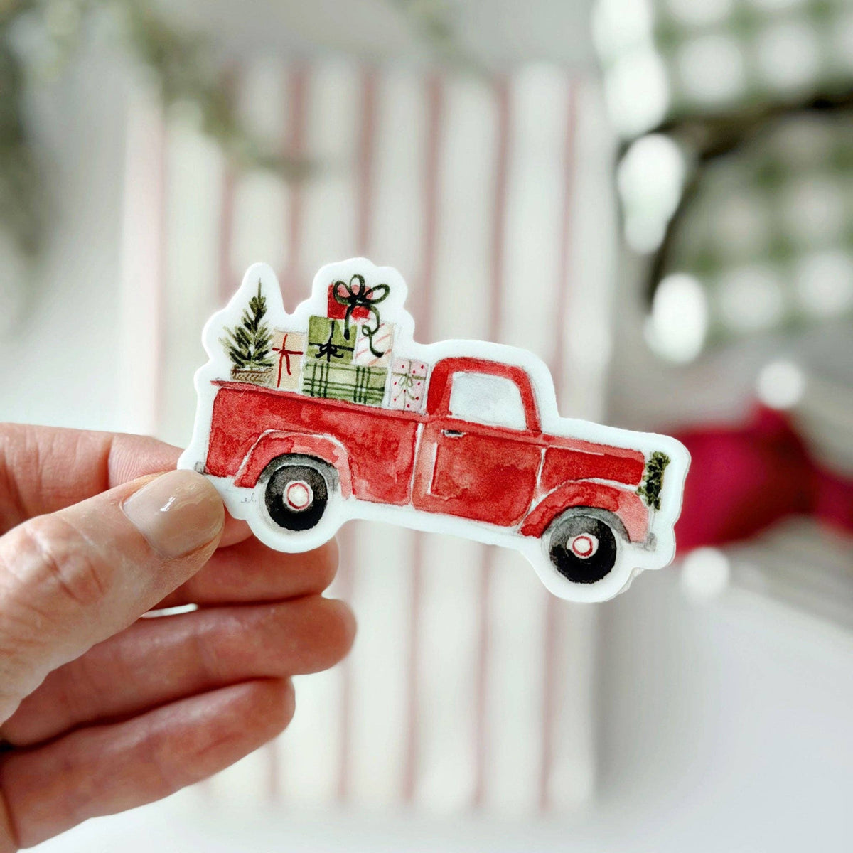 A hand holds a die-cut sticker featuring a red watercolor-style pickup truck carrying Christmas gifts and a small tree, against a softly blurred festive background.
