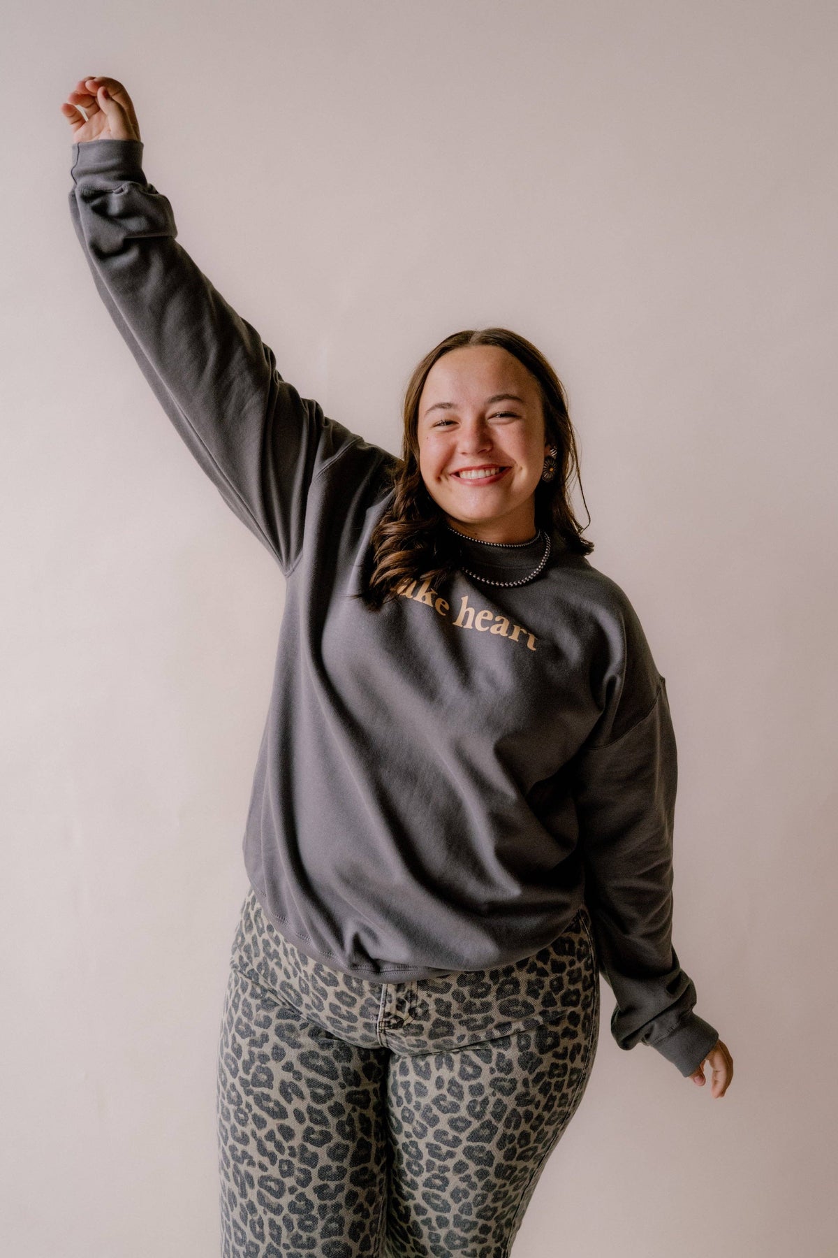 A woman posing in a dark gray sweatshirt with gold text that reads 'make heart' and leopard print leggings, standing against a plain light beige wall with one arm raised.