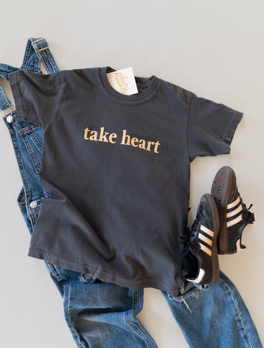 A dark charcoal gray graphic t-shirt with the words 'take heart' printed in a gold serif font, styled in a flat lay with blue denim overalls and black Adidas sneakers on a light gray background.