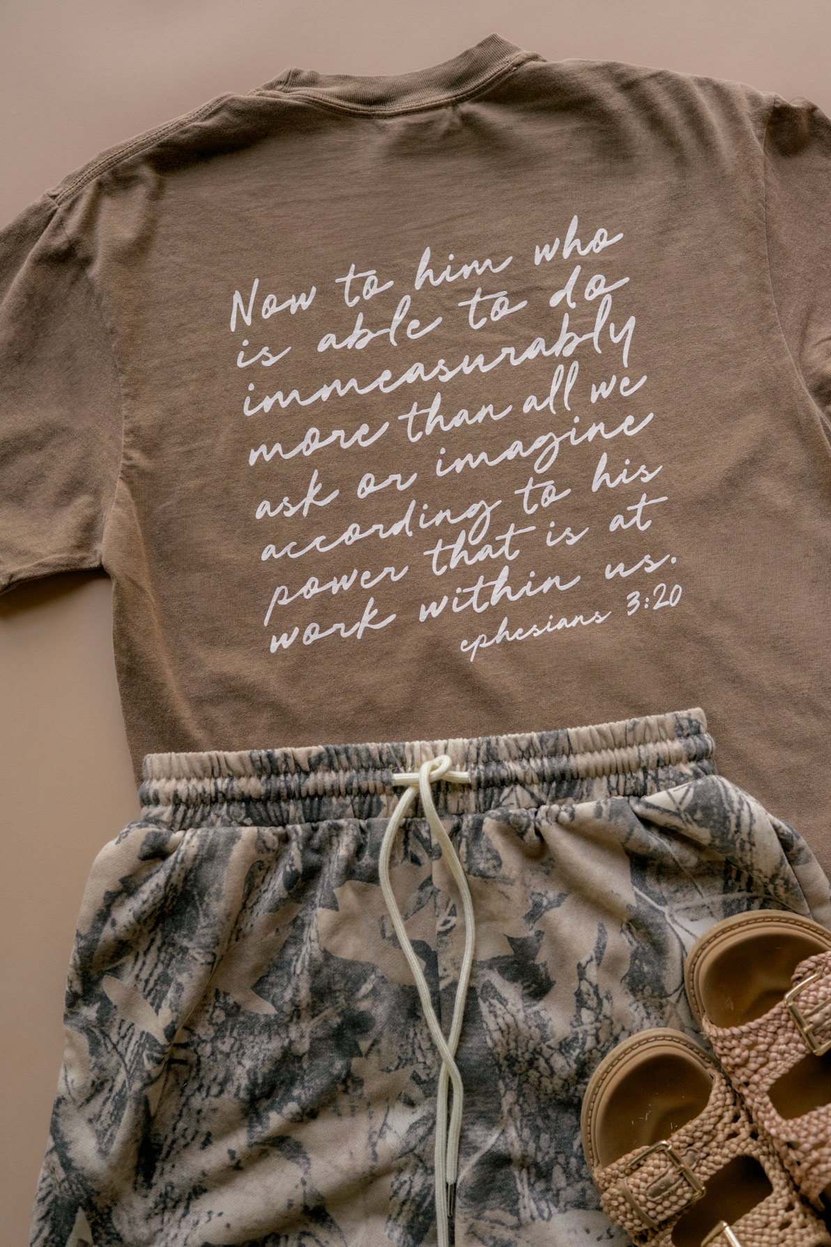A styled flat-lay of a taupe brown t-shirt with white script text, paired with a pair of gray and white patterned sweatpants and brown sandals on a solid beige background.