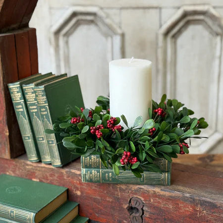 Decorative setup with a white candle, green wreath with red berries, and green books on a wooden surface.