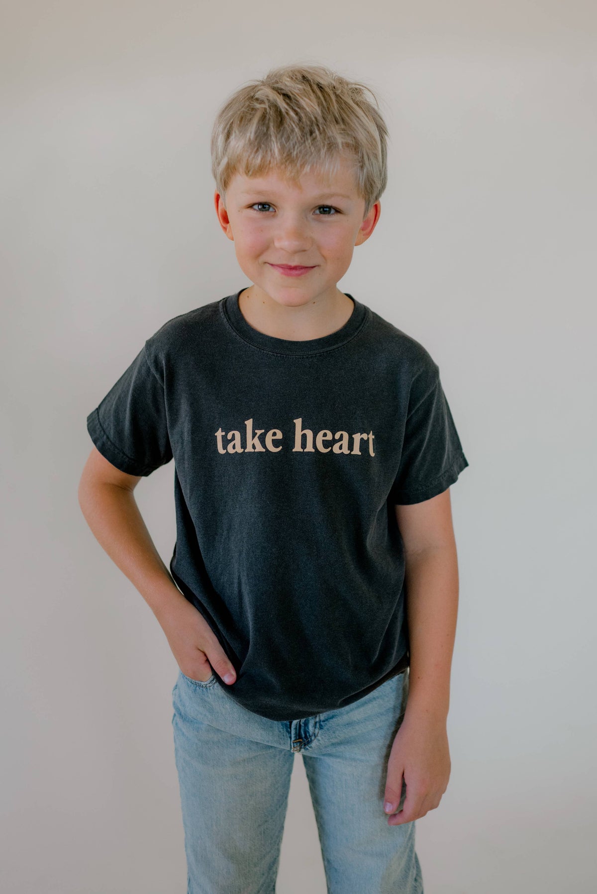 A young boy with blonde hair wearing a dark charcoal gray t-shirt with the words 'take heart' printed in a light beige gothic font, paired with light blue jeans, standing against a plain light gray background.