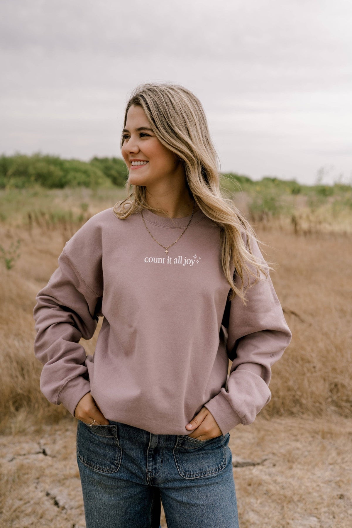 A woman wearing a dusty mauve crewneck sweatshirt with the text 'count it all joy' in white script, paired with blue jeans, standing outdoors in a dry grassy field under an overcast sky.