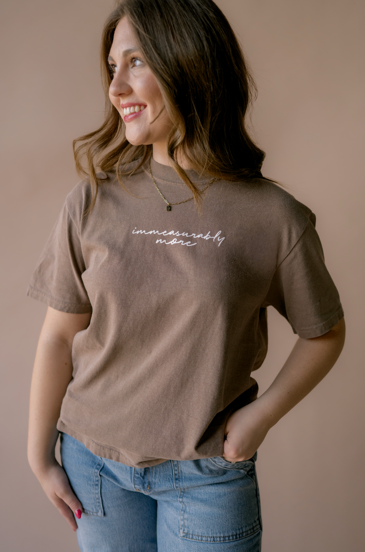 A woman wearing a brown t-shirt with white cursive text that reads 'immeasurably more' and blue jeans, standing against a solid beige background.