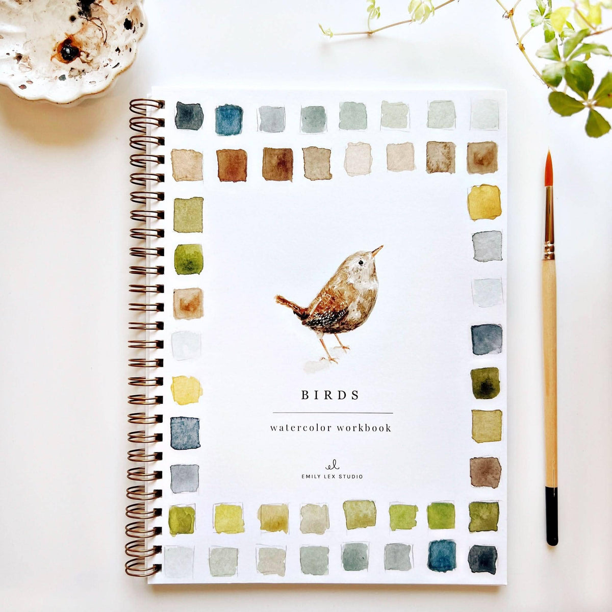 A spiral-bound watercolor workbook titled 'BIRDS' by Emily Lee Studio, featuring a watercolor bird illustration and a border of watercolor swatches, styled on a white surface with a paintbrush and a small dish.