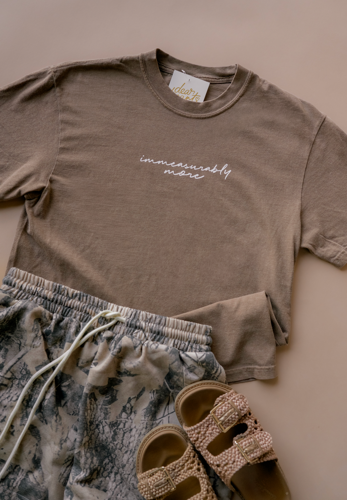 A styled flat-lay of a taupe brown t-shirt with 'immeasurably mere' printed in white script on the chest, paired with blue-gray patterned shorts and beige embellished slide sandals on a solid beige background.