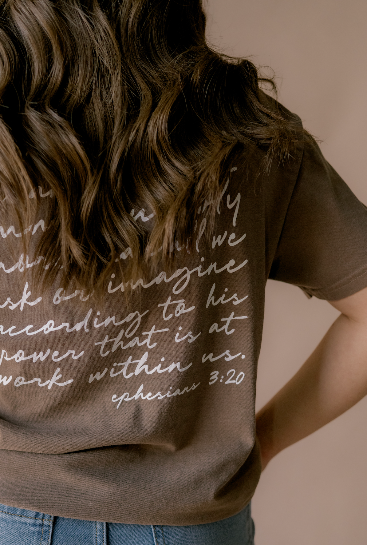 A person shown from behind wearing a dark brown t-shirt with white cursive text of a Bible verse, paired with blue jeans, against a solid beige background.