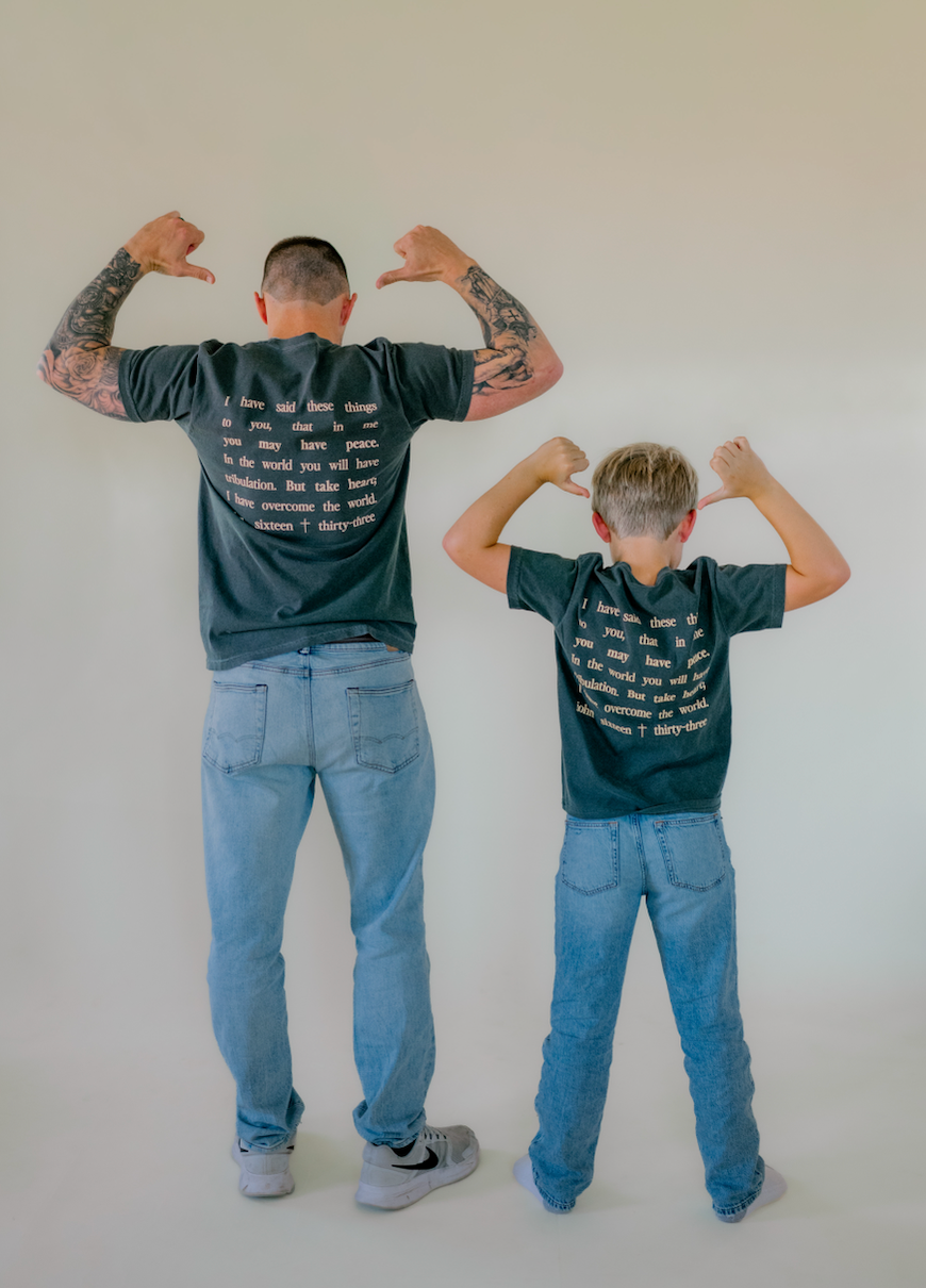 An adult man and a child standing side by side with their backs to the camera, both wearing matching dark grey graphic t-shirts and blue jeans, posing with their hands above their heads against a plain off-white background.