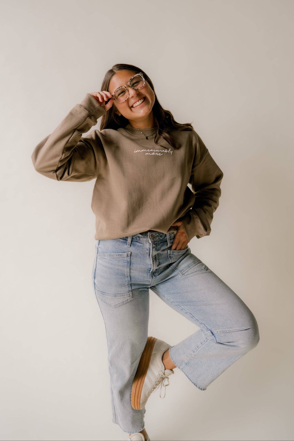 A woman posing in a studio setting wearing a taupe-brown crewneck sweatshirt with white script text on the chest, light-wash blue jeans, and white sneakers.