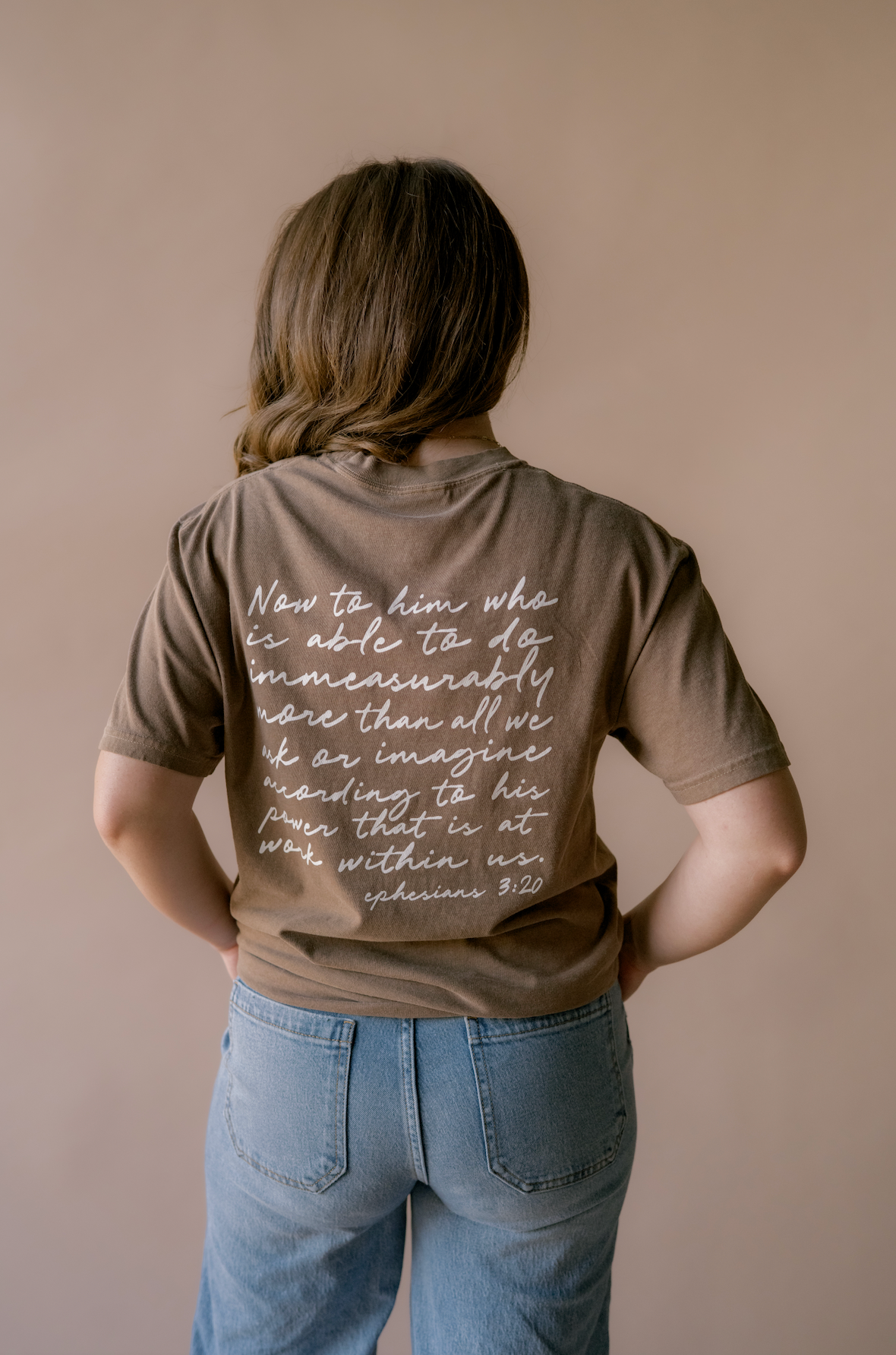 A person viewed from behind wearing a brown short-sleeve T-shirt with white cursive text printed on the back, paired with light blue jeans, against a plain beige wall.