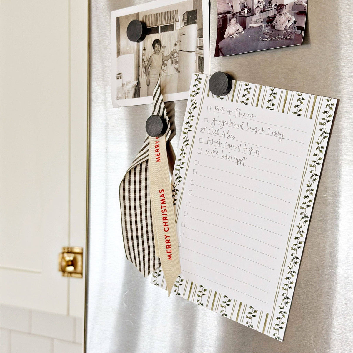 A stainless steel refrigerator door is shown with several magnets and papers attached: two small black-and-white photos, a striped and cream 'Merry Christmas' tag, and a lined notepad with a decorative border and handwritten checklist.