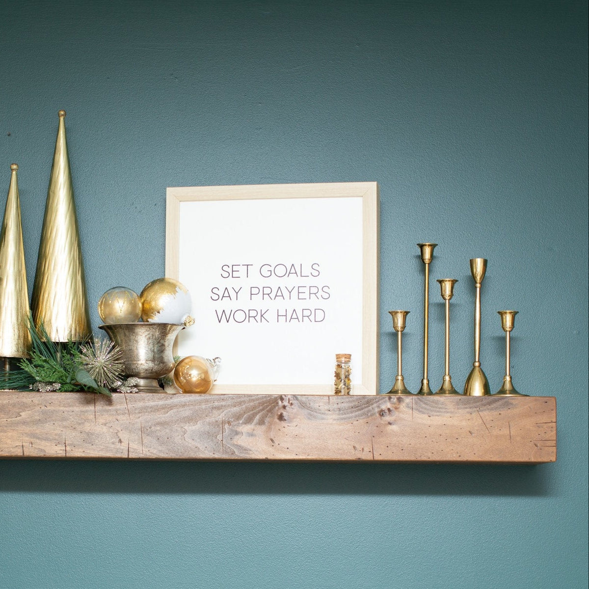 Decorative shelf with gold ornaments and candlesticks, greenery, and a framed quote against a teal wall.