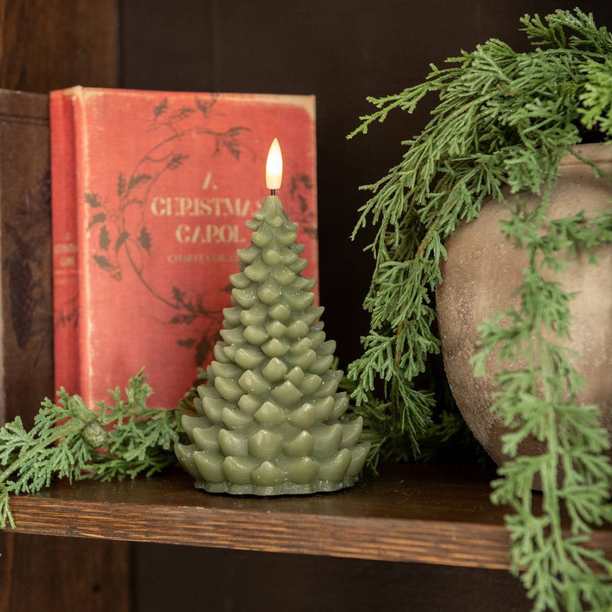 A lit Christmas tree candle is displayed on a wooden mantle with a vintage book and seasonal greens in a textured vase.