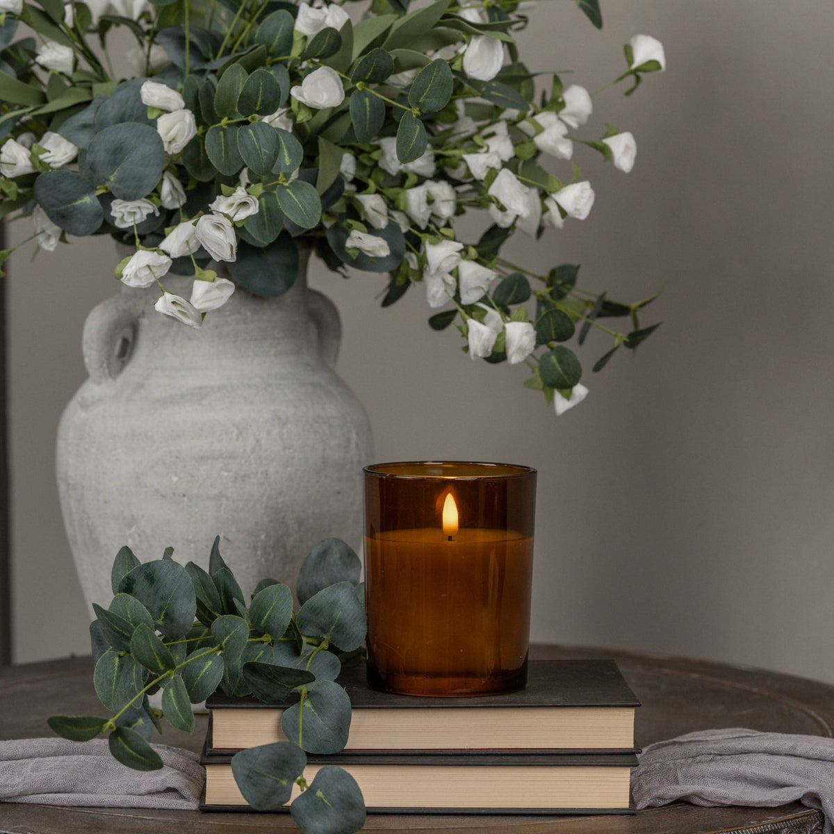 a Floral arrangement in a textured vase shown with a lit wick candle in amber vessel and a sprig of eucalyptus draped over books