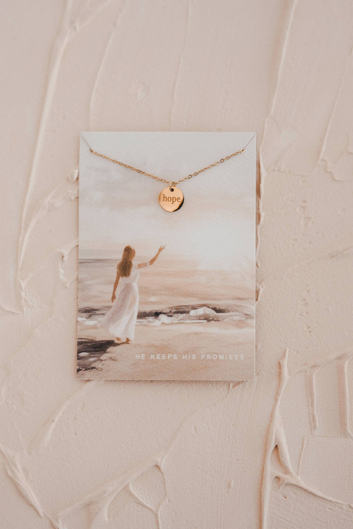 Gold necklace with 'hope' pendant on a beach scene card against a textured beige background
