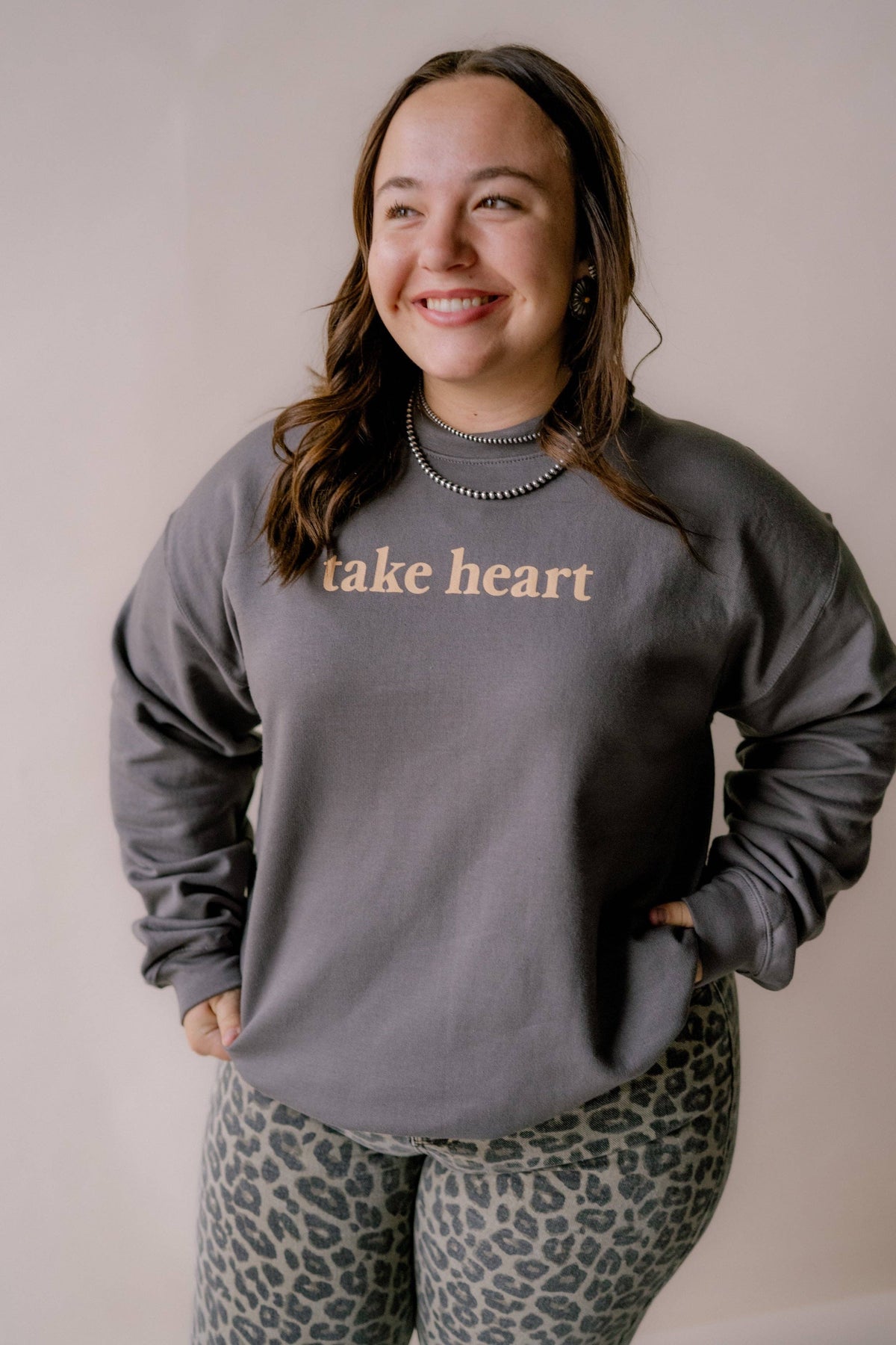 A woman wearing a dark gray crewneck sweatshirt with the words 'take heart' printed in a light beige font across the chest, paired with leopard print leggings, standing against a plain beige wall.