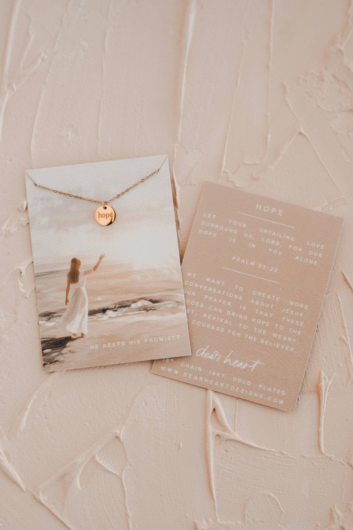 A gold necklace with a round engraved disc pendant displayed on a printed card next to a beige letter, both laid flat on a textured beige surface.