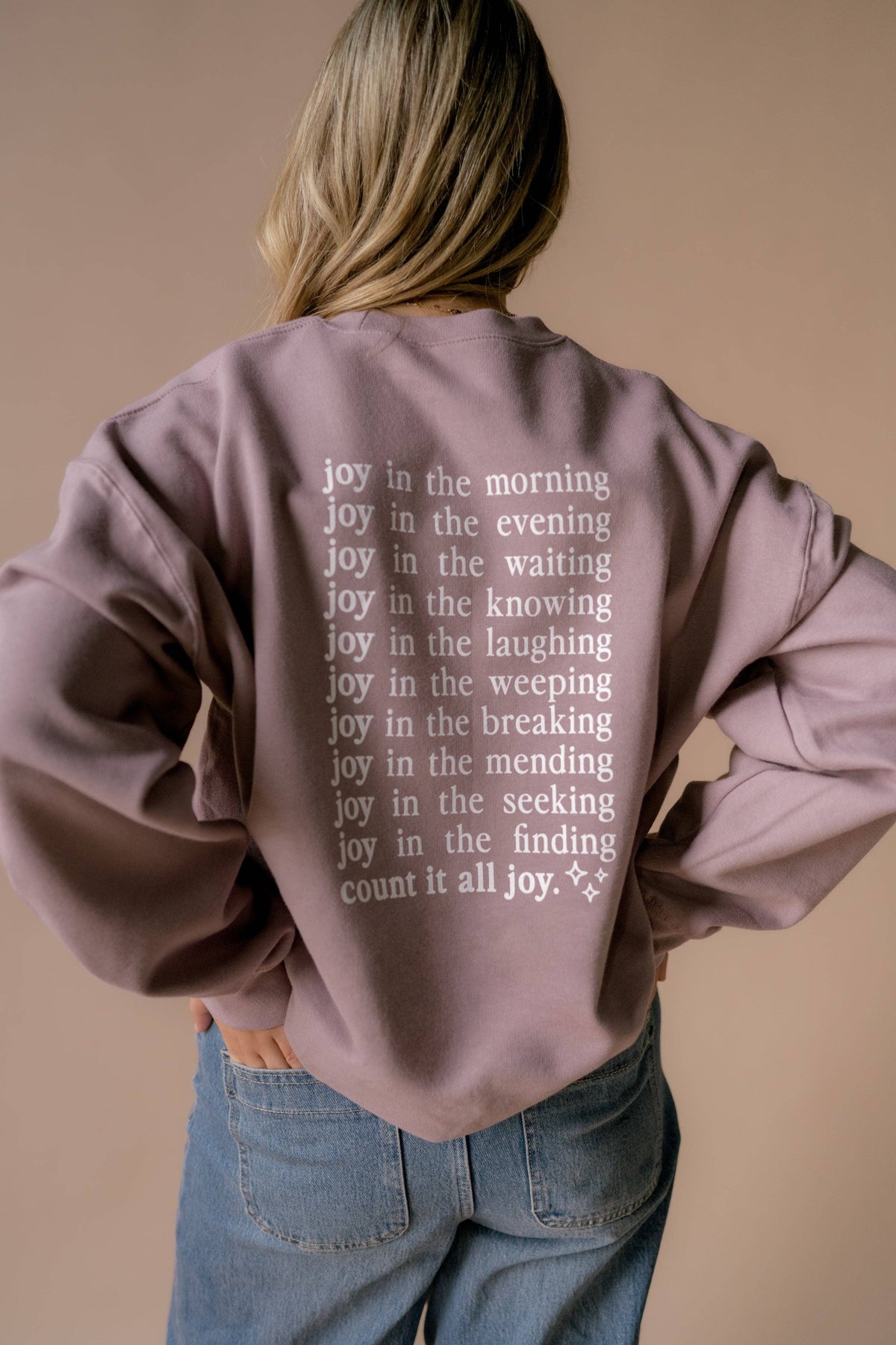 A woman viewed from behind wearing a mauve-purple sweatshirt with white printed text listing joys of daily life, paired with blue jeans against a solid beige background.