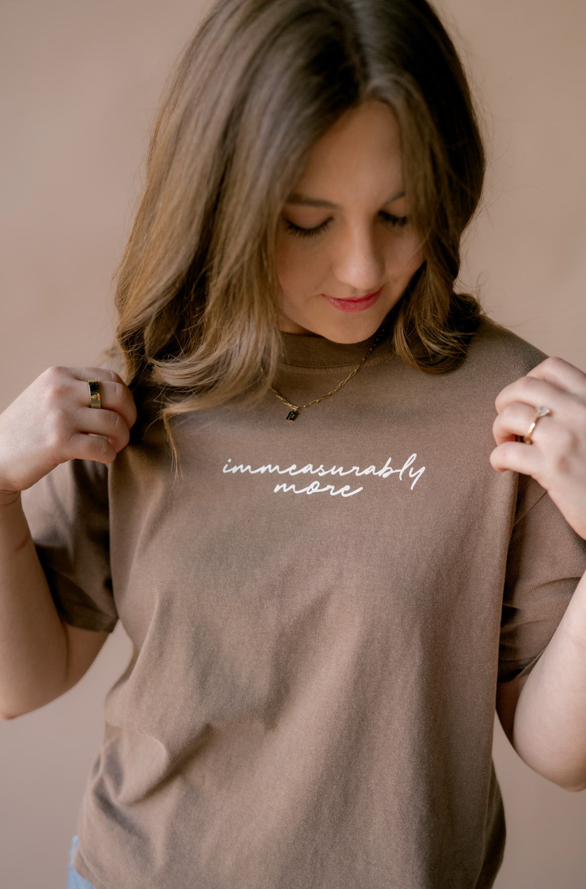 A woman wearing a taupe brown t-shirt with the phrase 'immeasurably more' printed in white script across the chest, posing against a solid beige background.