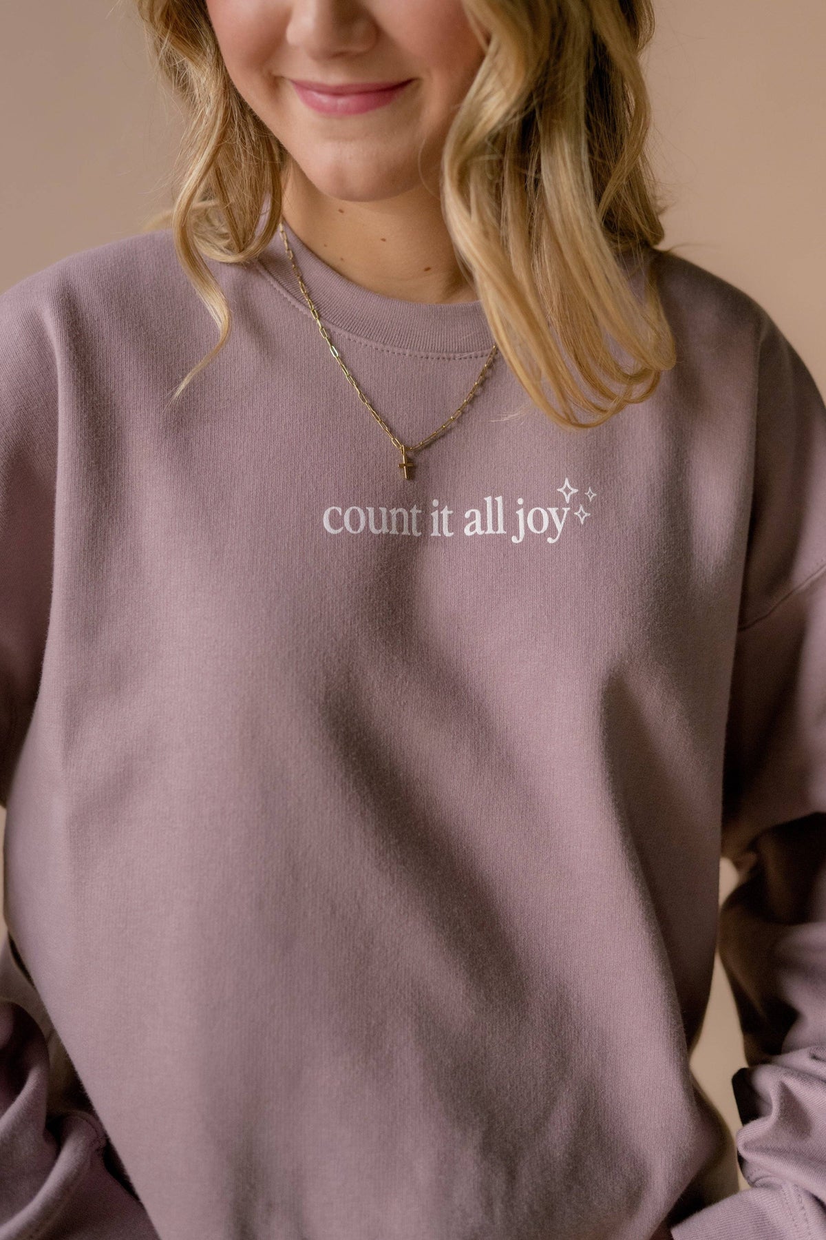 A close-up of a woman wearing a dusty mauve crewneck sweatshirt with the white printed text 'count it all joy' on the chest, accessorized with layered gold necklaces.