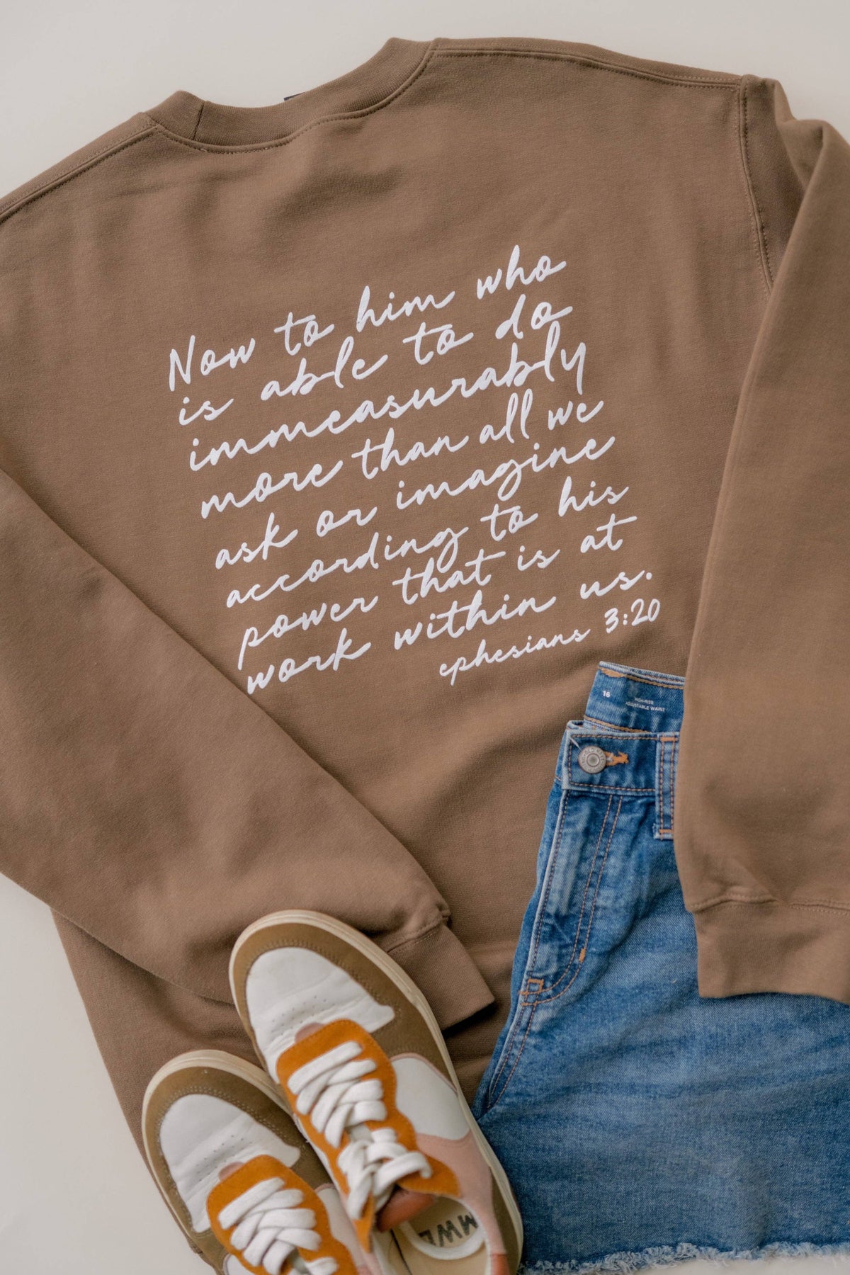 A flat-lay of a brown sweatshirt featuring a white printed bible verse from Ephesians 3:20 on the back. The sweatshirt is styled with blue denim jeans and white sneakers with orange accents on a light gray background.