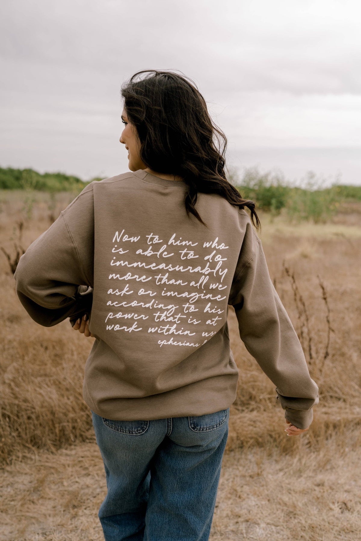 A woman viewed from behind wearing a taupe-brown sweatshirt with a white handwritten-style scripture printed on the back, standing in a dry grassy outdoor field under an overcast sky.
