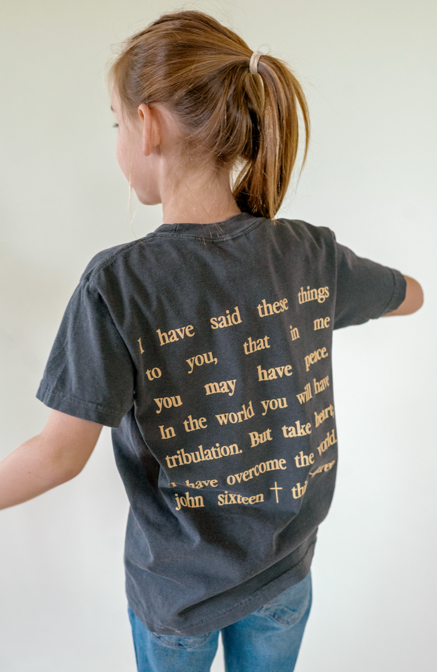 A child shown from the back wearing a faded black graphic t-shirt with gold text, paired with blue jeans, against a plain off-white background.
