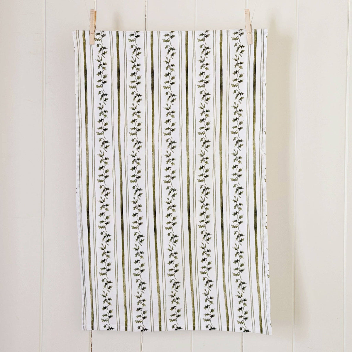 A rectangular fabric panel or curtain hanging from two wooden clothespins against a white paneled wall. The fabric has a vertical striped pattern with olive green and black leafy vine motifs on a white background.