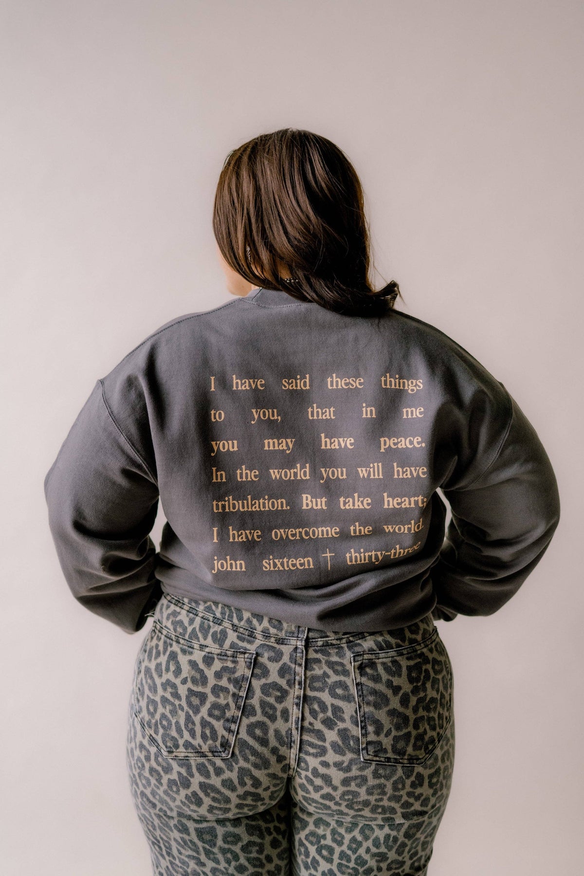 A person viewed from behind wearing a dark gray satin bomber jacket with gold text on the back, paired with leopard print pants, against a plain light gray background.