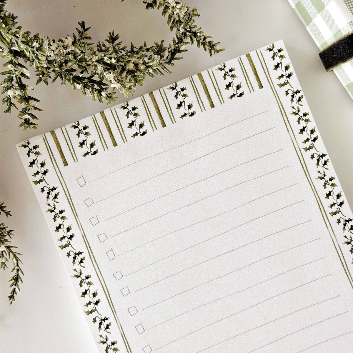 A lined notepad with a botanical border design, photographed from above on a light surface with decorative greenery and part of a green-and-white checkered item visible.