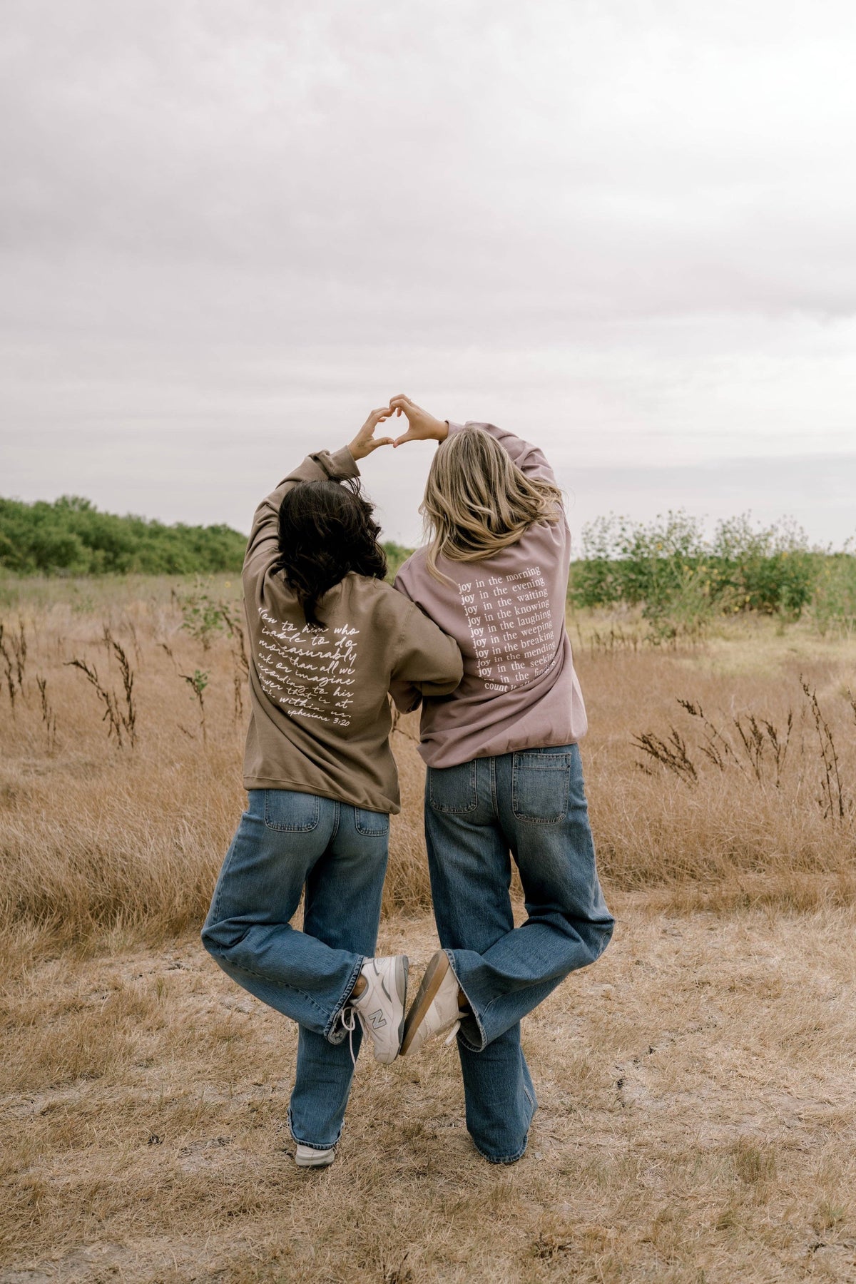 Two people standing in a dry grass field, viewed from behind, forming a heart shape with their hands over their heads. One wears a brown hoodie with white script text on the back and blue jeans, and the other wears a dusty pink hoodie with white text on the front and blue jeans.