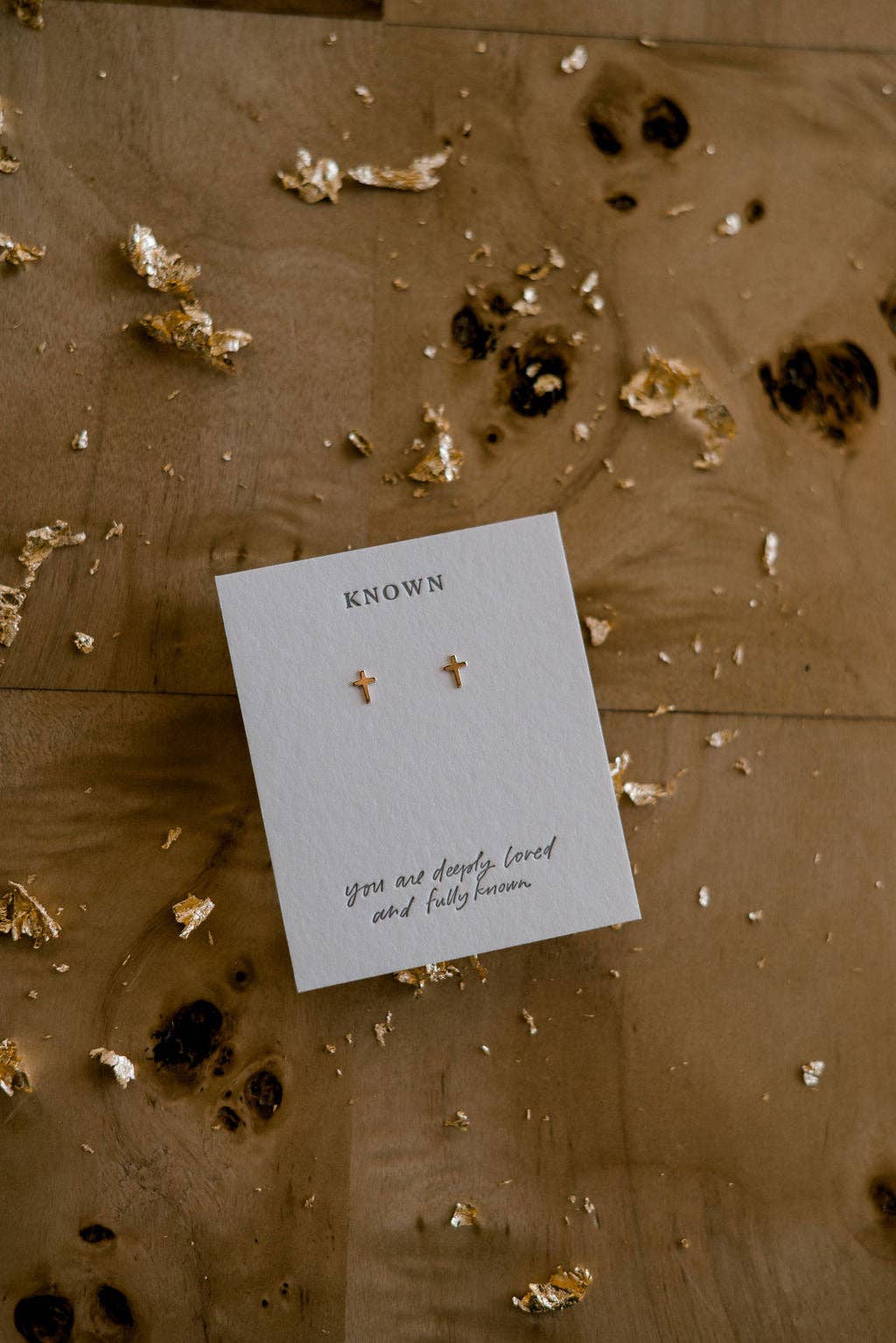 Pair of gold earrings on a 'Known' card with text on a wooden surface.
