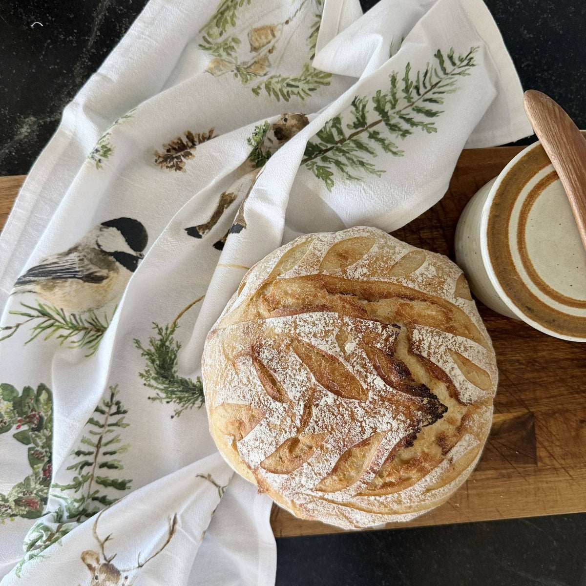 A round loaf of rustic bread sits on a wooden board next to a white tea towel printed with birds and green foliage, with a ceramic bowl partially visible.