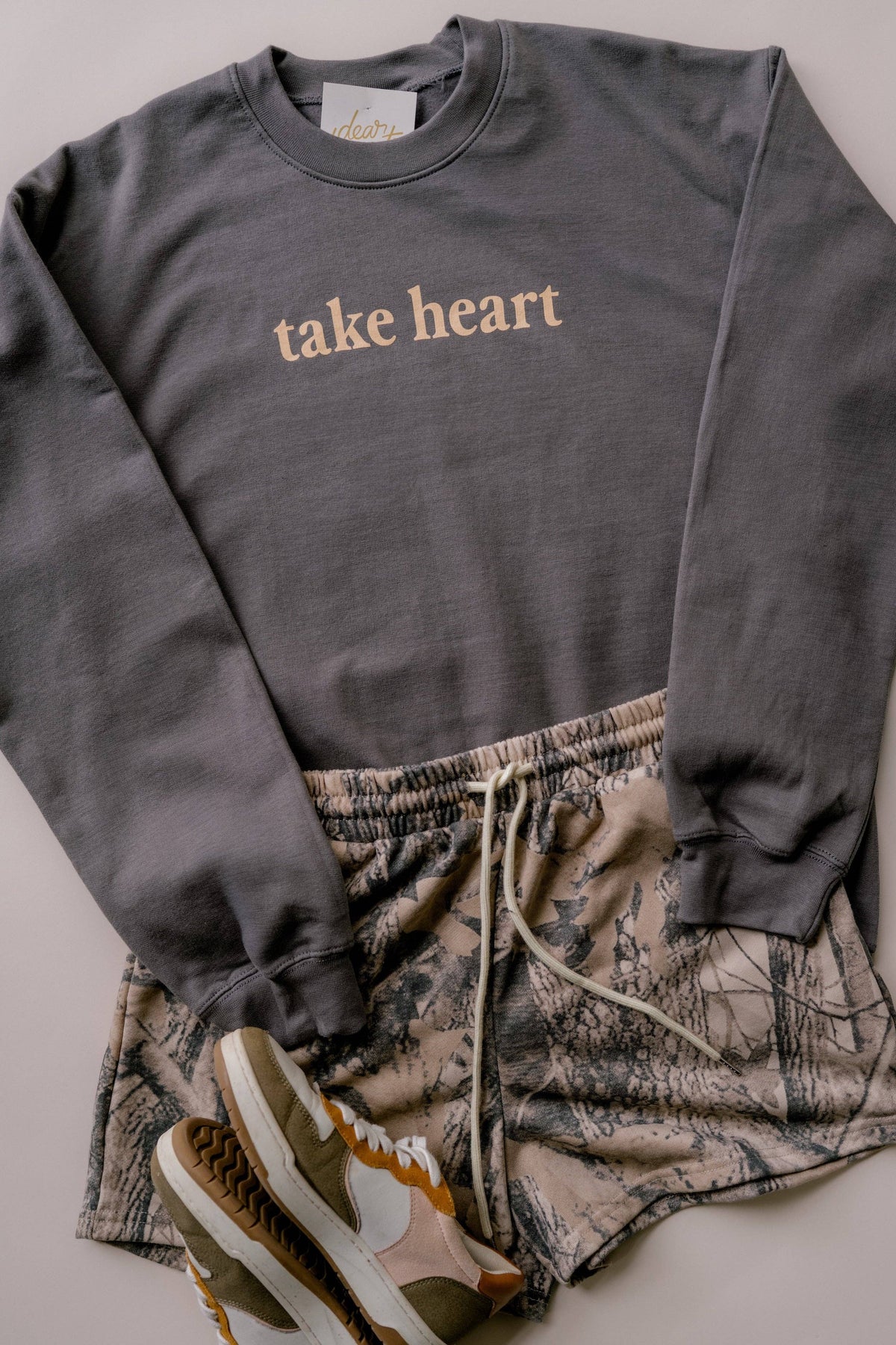 A dark gray crewneck sweatshirt with 'take heart' printed in beige across the chest, styled in a flat lay with camouflage drawstring shorts and olive sneakers on a neutral background.