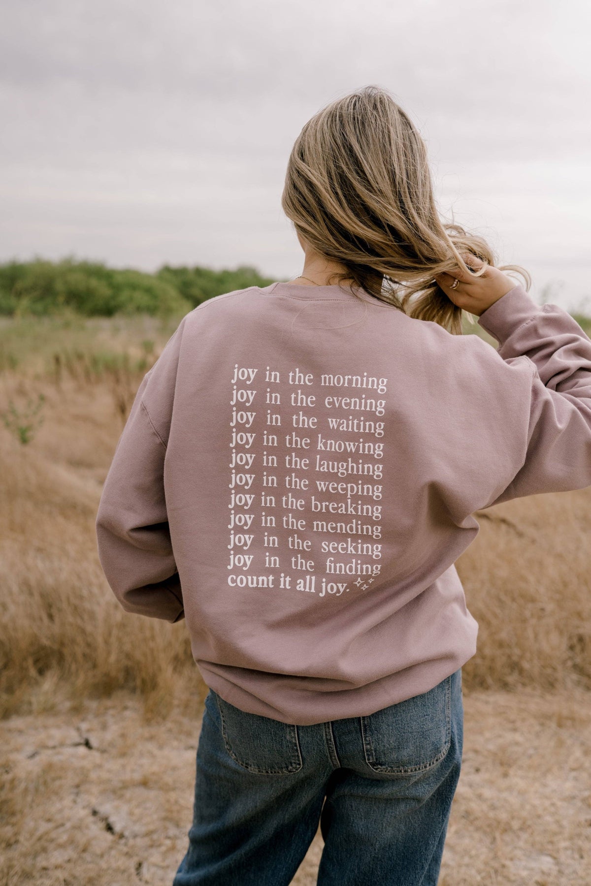 A woman viewed from behind wearing a dusty mauve sweatshirt with white printed text listing various emotions, paired with blue jeans in an outdoor field under an overcast sky.