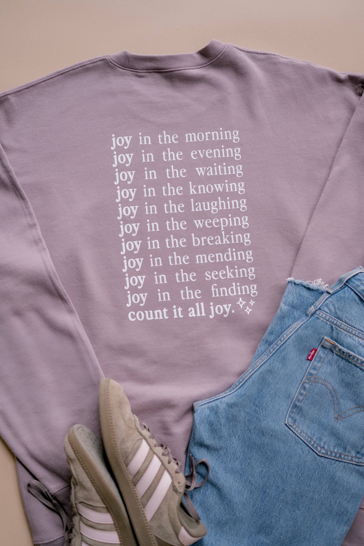 A lavender crewneck sweatshirt with white printed text listing joys of daily life, styled in a flat lay with blue jeans and beige sneakers on a beige background.