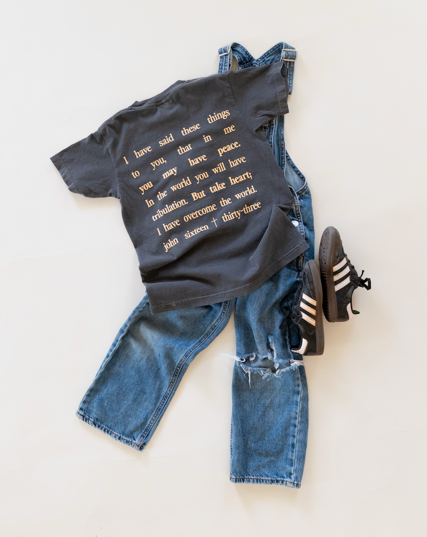 A styled flat-lay of a black graphic t-shirt with gold text, paired with blue denim overalls and black sneakers with white stripes, on a light cream background.