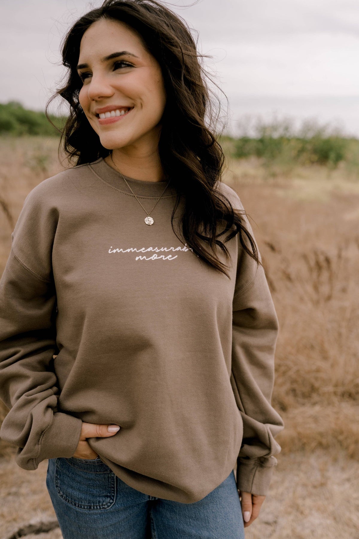 A woman wearing a taupe brown crewneck sweatshirt with white script text reading 'immeasurably more' on the chest, paired with blue jeans, standing outdoors in a dry grassy field.
