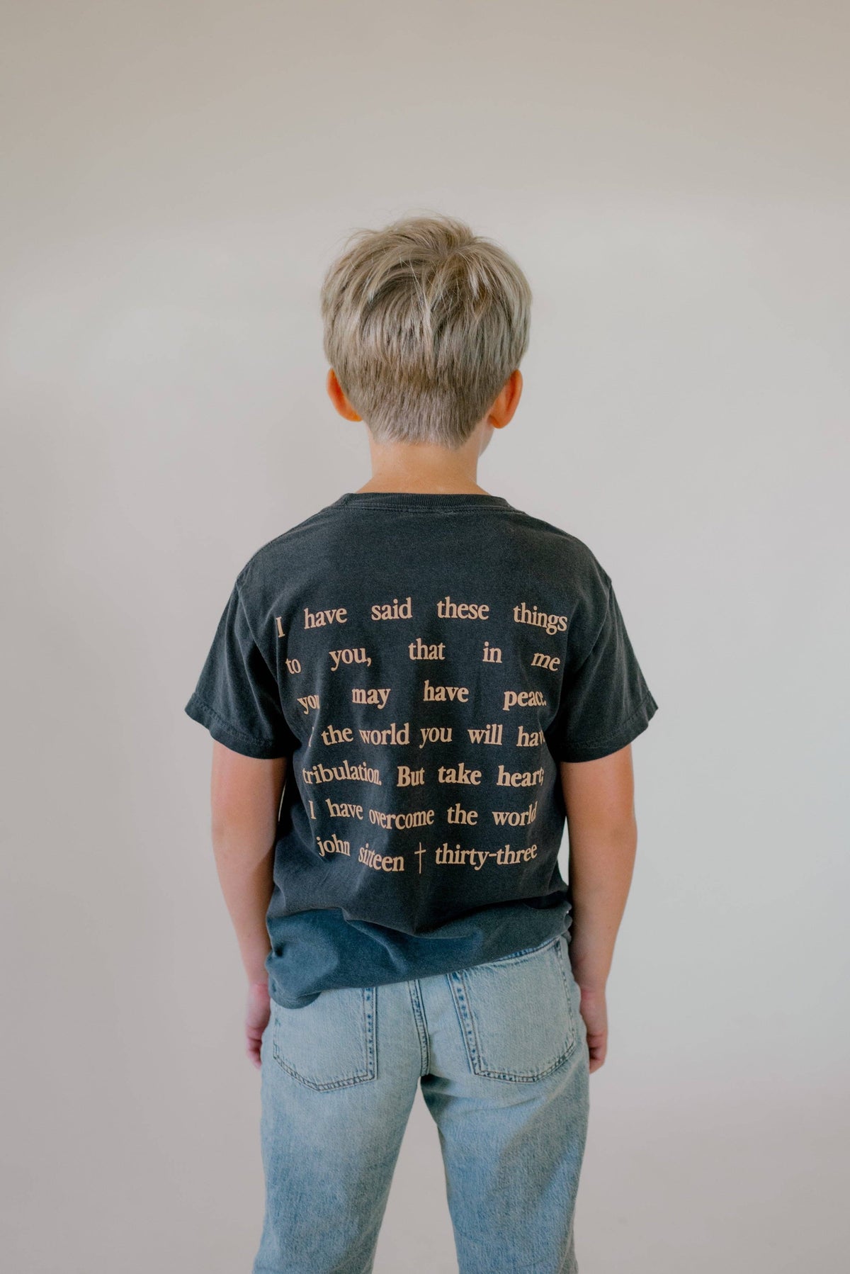 A child viewed from behind wearing a faded black graphic t-shirt with gold text and light wash blue jeans, standing against a plain light gray wall.