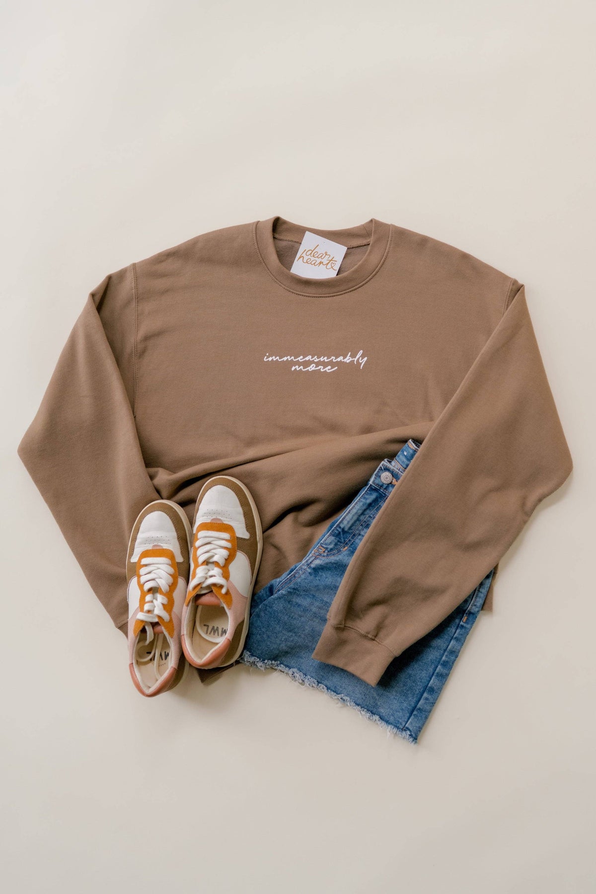 A styled flat-lay of a taupe crewneck sweatshirt with white script embroidery reading 'immeasurably  more'. The sweatshirt is layered over blue denim shorts, with white and orange sneakers placed at the bottom left, all on a light beige background.